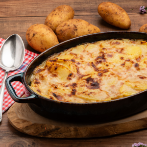 Überbackener Kartoffelauflauf in einer schwarzen Auflaufform, daneben rohe Kartoffeln und ein Löffel